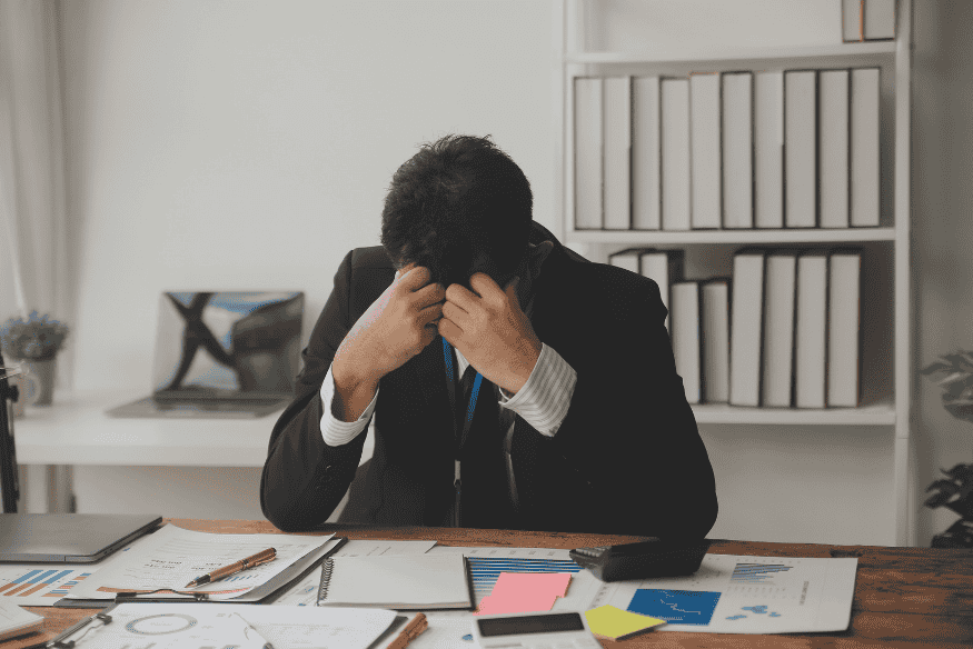 Ein gestresster Geschäftsmann sitzt an einem Schreibtisch mit Finanzunterlagen, hält den Kopf in den Händen und wirkt überfordert von seiner Arbeit.