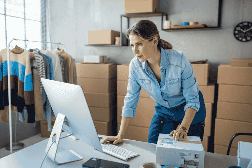 Eine Frau arbeitet in einem kleinen Büro oder Lager, steht vor einem Computer und bereitet Pakete für den Versand vor.
