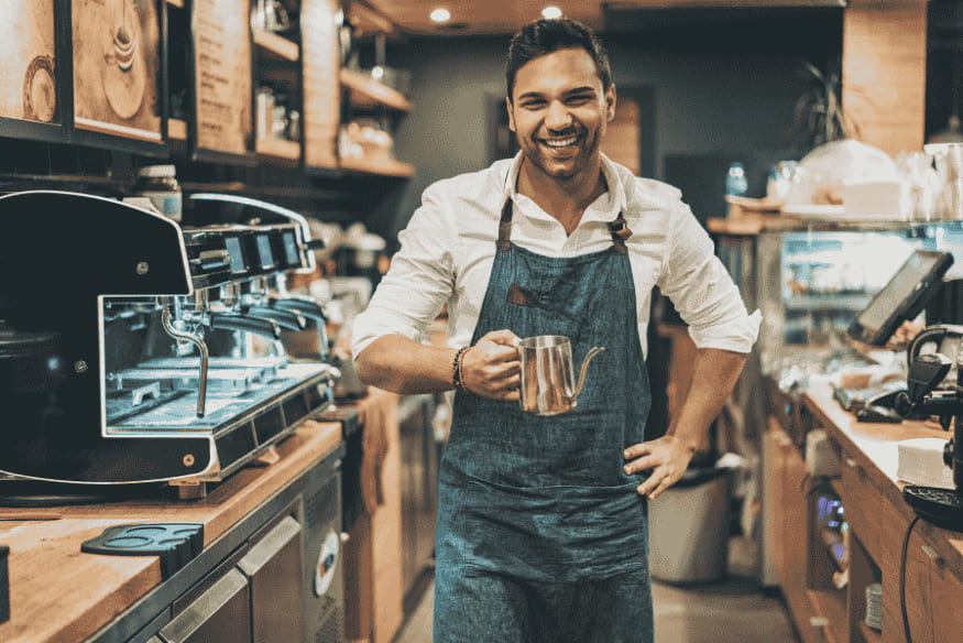 Ein lächelnder Barista steht hinter der Theke eines Cafés, hält eine Milchkanne in der Hand und trägt eine Schürze.