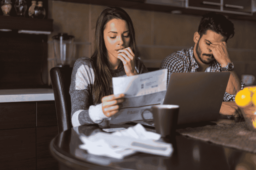 Ein besorgtes Paar sitzt am Tisch mit Rechnungen und Laptop, während es über finanzielle Unterlagen nachdenkt.
