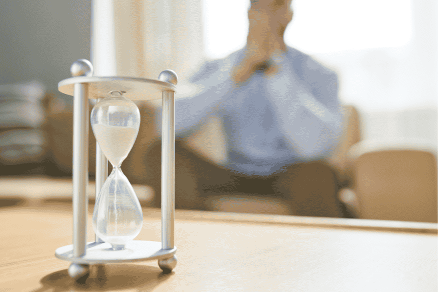 Nahaufnahme einer stilvollen Sanduhr auf einem Tisch, mit einem nachdenklichen Mann im Hintergrund – symbolisiert die 7-Tage-Regel für bessere finanzielle Entscheidungen.