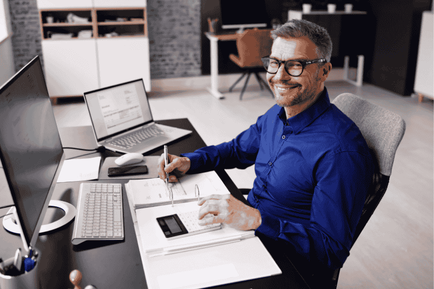 Lächelnder Mann am Schreibtisch mit Laptop, Taschenrechner und Finanzunterlagen – symbolisiert die strukturierte Übersicht über alle aktiven Bankkonten für mehr finanzielle Kontrolle.
