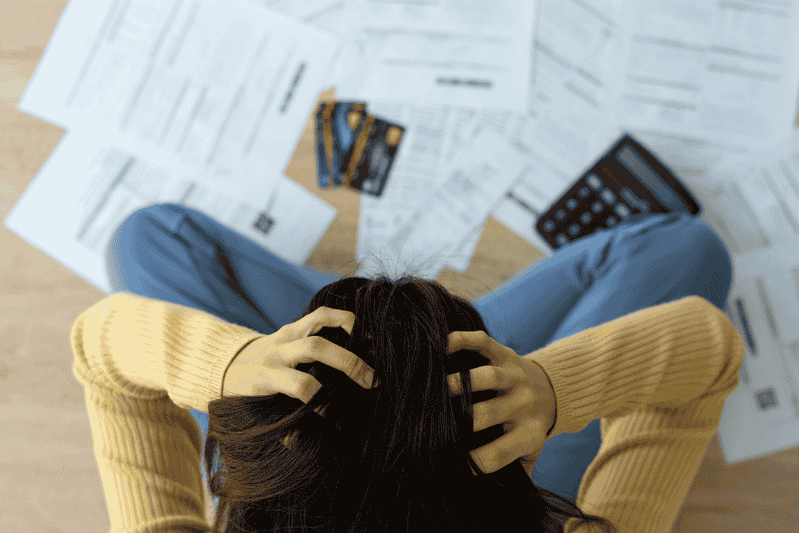 Eine Frau sitzt auf dem Boden, hält verzweifelt den Kopf in den Händen, umgeben von Rechnungen, Kreditkarten und einem Taschenrechner.