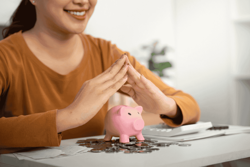 Eine lächelnde Frau sitzt am Tisch, bildet mit den Händen ein Dach über einem Sparschwein, umgeben von Münzen und Papieren.