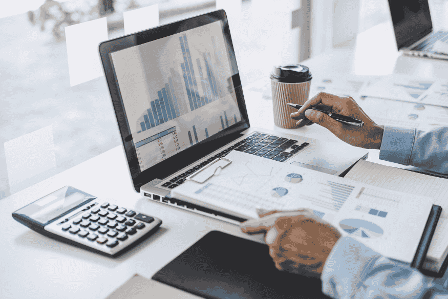 Ein Laptop zeigt Finanzdiagramme und Balkengrafiken, daneben liegen Taschenrechner, Notizen und eine Kaffeetasse auf einem Schreibtisch.