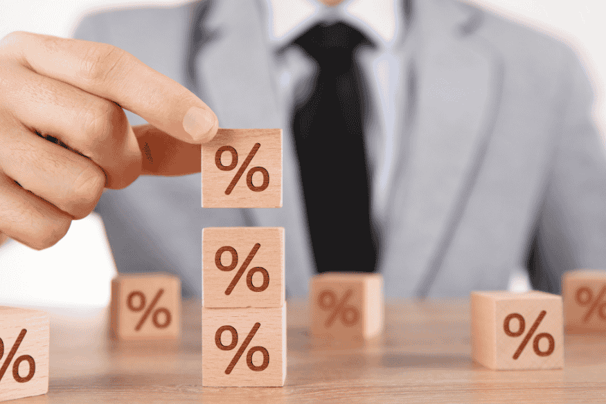 Ein Mann im Anzug stapelt Holzklötze mit Prozentzeichen – Symbol für Zinsentwicklung und Ertragssteigerung.
