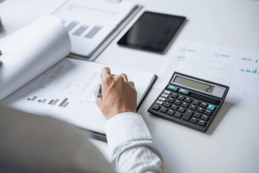 Eine Person arbeitet mit Finanzunterlagen, Diagrammen und Taschenrechner auf einem Schreibtisch, während sie Zahlen überprüft und Notizen macht.