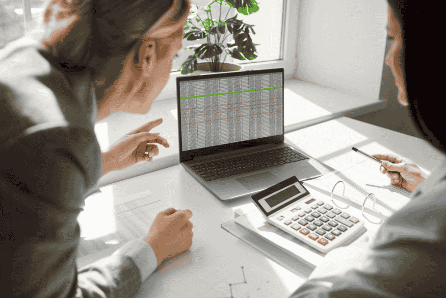 Zwei Personen analysieren Finanzdaten auf einem Laptop, besprechen Zahlen und nutzen Taschenrechner sowie Dokumente zur Entscheidungsfindung.