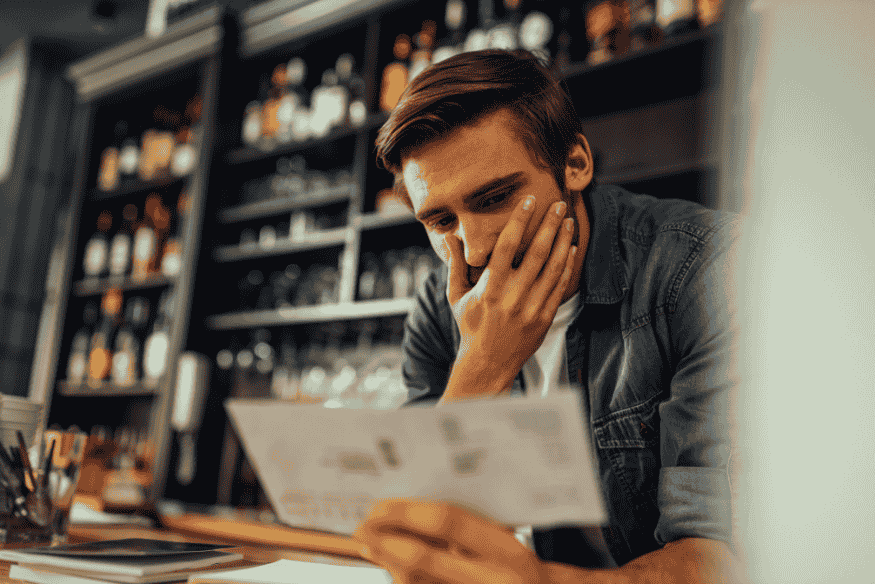 Ein besorgter Mann sitzt in einem Café oder Geschäft und betrachtet nachdenklich ein Dokument, vermutlich eine Rechnung oder einen Steuerbescheid.