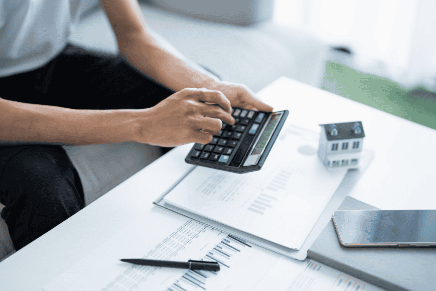 Eine Person berechnet finanzielle Daten mit einem Taschenrechner, auf dem Tisch liegen Rechnungsunterlagen, ein Smartphone und ein kleines Modellhaus.