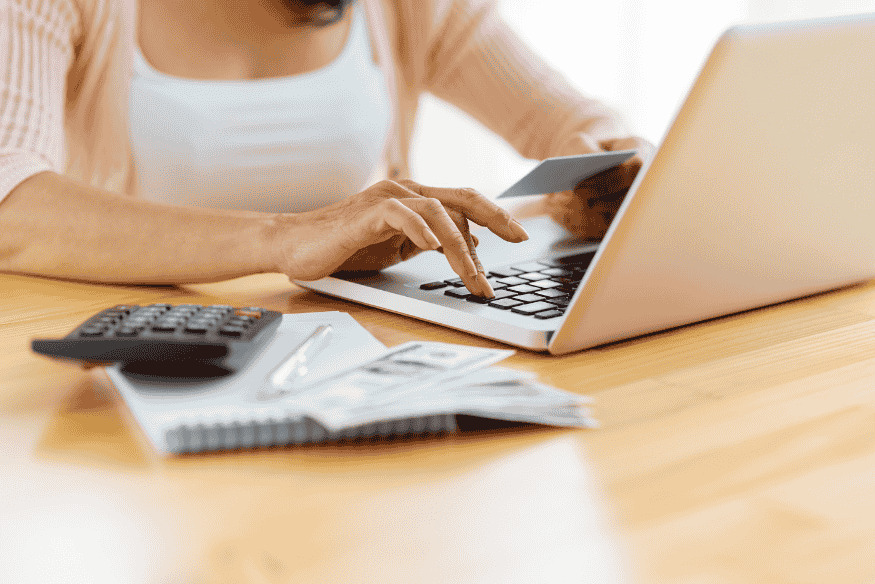 Eine Person gibt Zahlungsdaten am Laptop ein, hält eine Kreditkarte in der Hand und hat einen Taschenrechner sowie Rechnungen auf dem Tisch liegen.