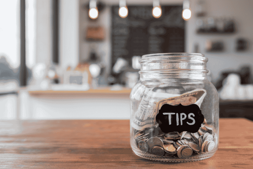 Ein Glas mit der Aufschrift „Tips“ steht auf einem Holztisch und ist mit Münzen und Geldscheinen gefüllt.