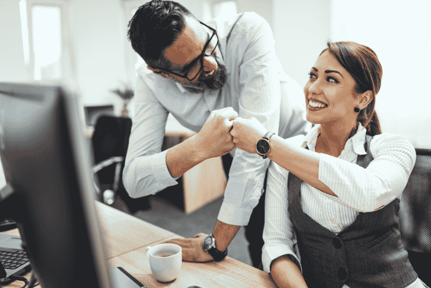Zwei Kollegen im Büro lächeln und geben sich einen freundschaftlichen Faustgruß, während sie an einem Schreibtisch sitzen.