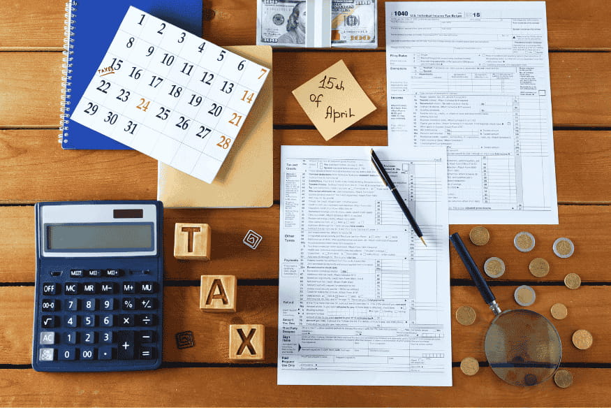 Auf einem Holztisch liegen Steuerformulare, ein Kalender mit markiertem Termin, ein Taschenrechner, Münzen, Geldscheine und Holzblöcke mit der Aufschrift „TAX“.