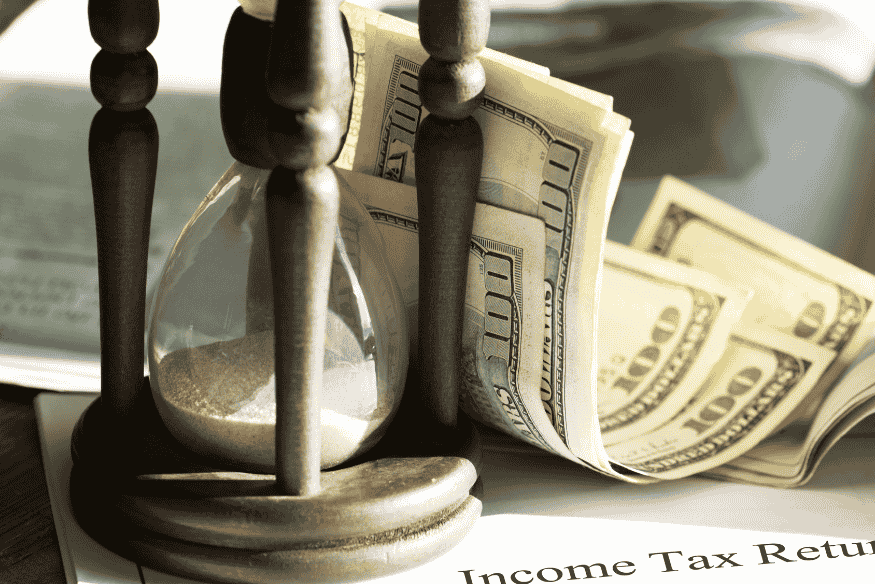 Ein Stapel Geldscheine liegt neben einer Sanduhr auf Steuerdokumenten – Symbol für den Zusammenhang zwischen Zeit, Geld und steuerlichen Fristen.