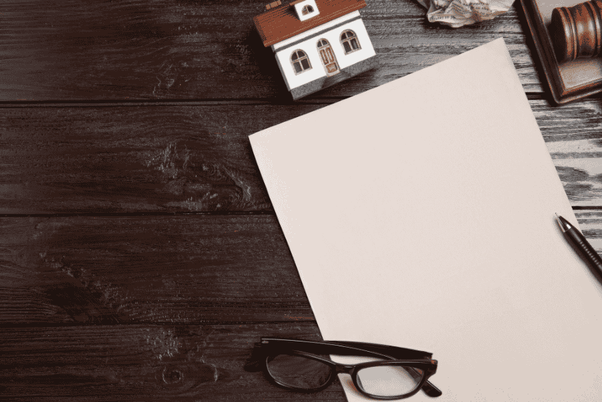 Auf einem Holztisch liegen eine leere Seite Papier, ein Stift, eine Brille und ein kleines Modellhaus – Symbol für finanzielle Planung und Vermögensaufbau.