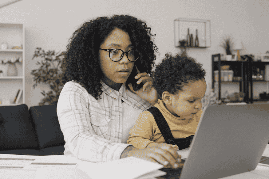 Mutter arbeitet am Laptop mit Kleinkind auf dem Schoß und organisiert Finanzen in der Elternzeit